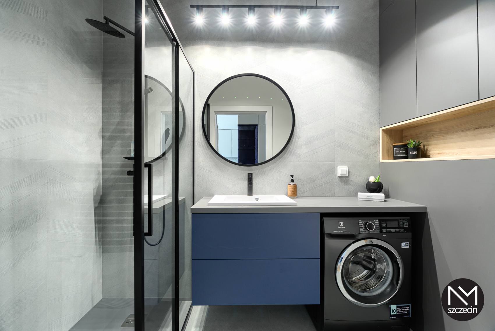Nowoczesna łazienka w szarościach z akcentami granatu: kabina prysznicowa typu walk-in, okrągłe lustro nad umywalką z szafką, pralka w zabudowie, oświetlenie punktowe.
