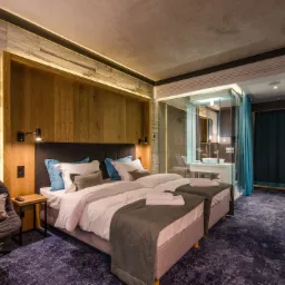 Nowoczesny pokój hotelowy z dwoma łóżkami, drewnianą ścianą z podświetleniem LED, szarą tapetą imitującą beton i przeszkloną łazienką.