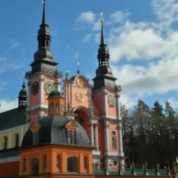 Barokowa fasada kościoła z dwoma wieżami zegarowymi, pomalowana na odcienie różu i pomarańczy, z kopułą i rzeźbami na tle błękitnego nieba z chmurami.