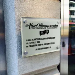 Tabliczka informacyjna z nazwą firmy tłumaczeniowej Alan Tłumaczenia, umieszczona na szorstkiej, jasnej ścianie obok metalowych drzwi wejściowych. Widoczne dane kontaktowe: adres e-mail, numer...