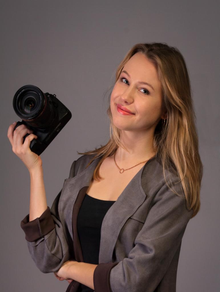 Uśmiechnięta blondynka trzymająca aparat fotograficzny z obiektywem, ubrana w szarą marynarkę na tle szarego tła.