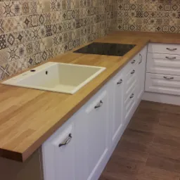 Jasna kuchnia z drewnianym blatem, wzorzystą mozaiką nad blatem i brązowymi płytkami podłogowymi imitującymi drewno.