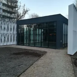 Nowoczesny pawilon z przeszklonymi ścianami i dekoracyjnymi, ażurowymi panelami, otoczony gruntem i ścieżką z kamyczków.