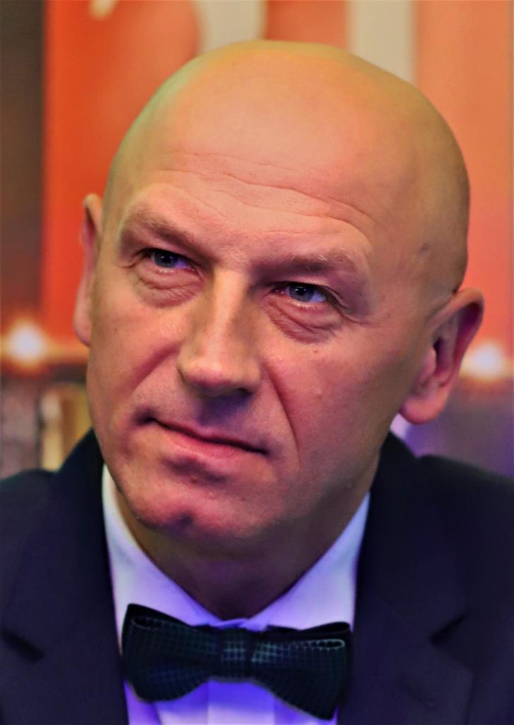 Portret mężczyzny z łysą głową, w ciemnym garniturze i muszce w kratę, zamyślony wyraz twarzy, oświetlenie sceniczne.