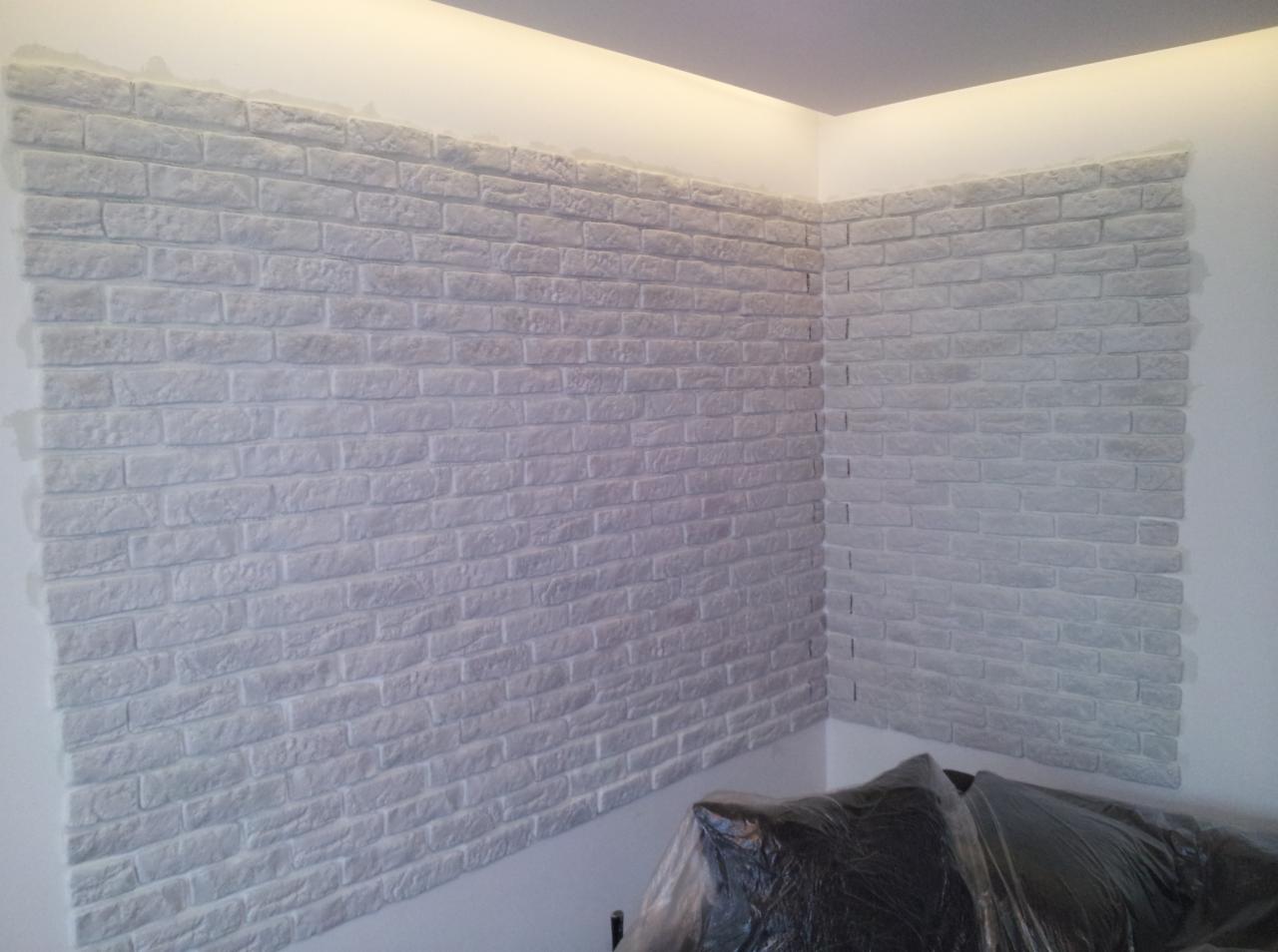 Ściana wykończona białymi płytkami imitującymi cegłę, widoczne nierówności fug, fragment mebla zabezpieczonego folią w dolnej części kadru.