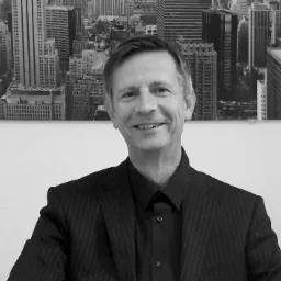 Czarno-białe portretowe zdjęcie uśmiechniętego mężczyzny w garniturze w paski, na tle ściany z fototapetą przedstawiającą panoramę miasta.