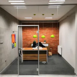 Szklane drzwi biurowe z metalową ramą, widok na wnętrze z ceglaną ścianą, drewnianym biurkiem i trzema zielonymi lampami wiszącymi.