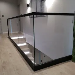 Nowoczesna szklana barierka z czarną ramą zabezpieczająca schody z podświetleniem LED, widok z góry na drewnianą podłogę.