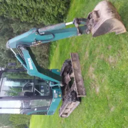 Turkusowa mini koparka marki Zeppelin, stojąca na trawie, z łyżką skierowaną w górę, widoczna kabina operatora i gąsienice.