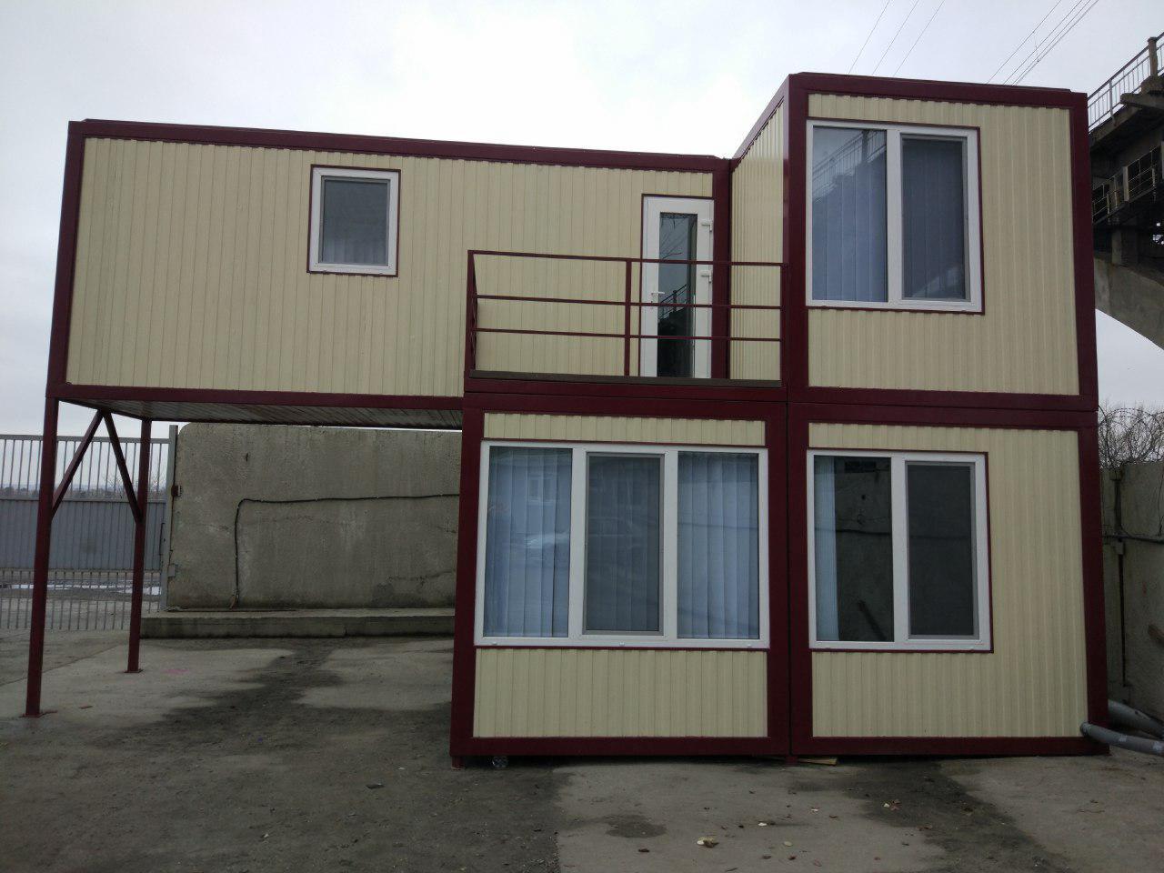 Dwukondygnacyjny dom modułowy w Poznaniu, złożony z kontenerów, z balkonem na piętrze, na tle betonowego muru i pochmurnego nieba.