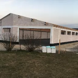 Budynek gospodarczy w trakcie budowy z bloczków, widoczne ocieplenie fundamentów i przygotowane drewno na więźbę dachową, na pierwszym planie krzewy bez liści.