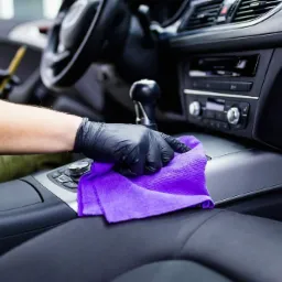 Wnętrze samochodu czyszczone fioletową ściereczką z mikrofibry przez osobę w czarnych rękawiczkach, widoczne elementy deski rozdzielczej, tunelu środkowego i fotela kierowcy.