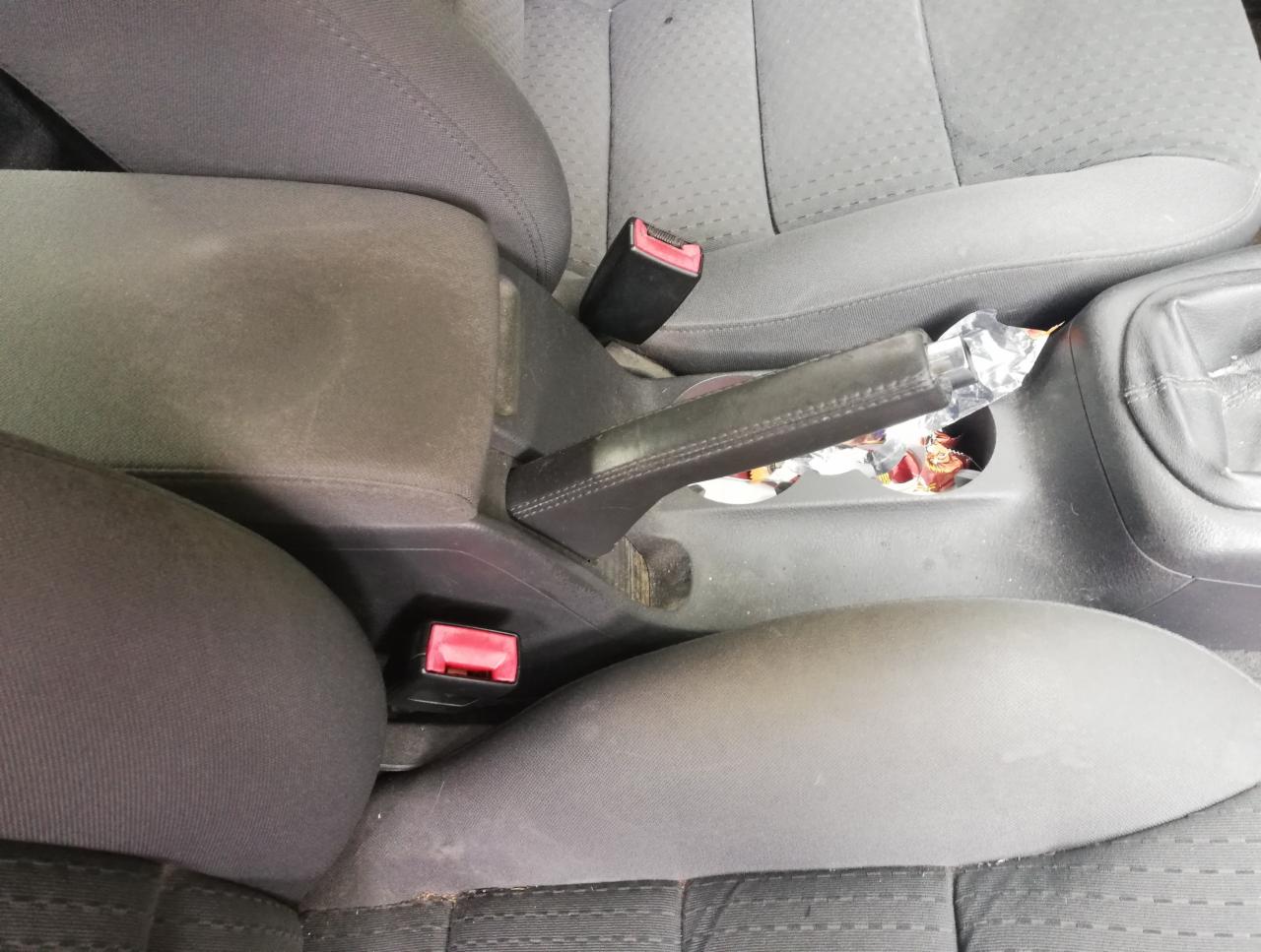 Brudne, szare wnętrze samochodu z widocznym podłokietnikiem, dźwignią hamulca ręcznego i resztkami jedzenia w uchwycie na kubek.