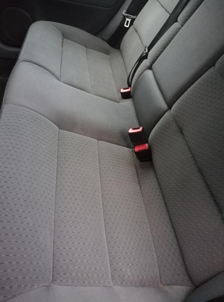 Szare, tekstylne siedzenie samochodowe z widocznymi pasami bezpieczeństwa i zagłówkami; materiał fotela wydaje się być lekko zabrudzony.