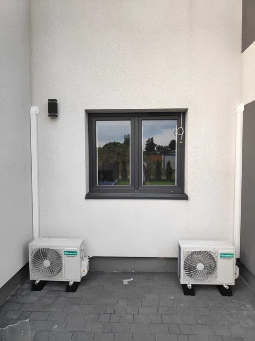 Dwa identyczne klimatyzatory Hisense umieszczone symetrycznie pod oknem na zewnątrz budynku z teksturowaną białą elewacją.