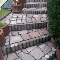 Schody zewnętrzne wyłożone nieregularnymi płytami kamiennymi, obrzeża z betonowych elementów cylindrycznych, donice z roślinami i figurką sowy, widok z dołu.