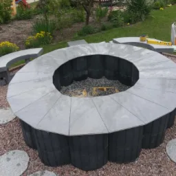 Okrągłe palenisko ogrodowe z szarej kostki betonowej i kamiennych płyt, otoczone żwirem i elementami małej architektury w ogrodzie.