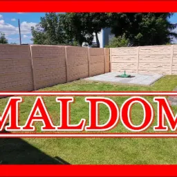 Nowoczesny, beżowy płot betonowy z dwoma różnymi wzorami paneli, otaczający trawnik z wyłożonymi płytami i zielonym słupkiem pośrodku, w słoneczny dzień.