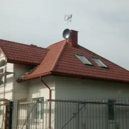 Antena satelitarna i antena naziemna zamontowane na dachu domu z czerwoną dachówką, widok zza ogrodzenia z metalowych prętów, rusztowanie przy ścianie.