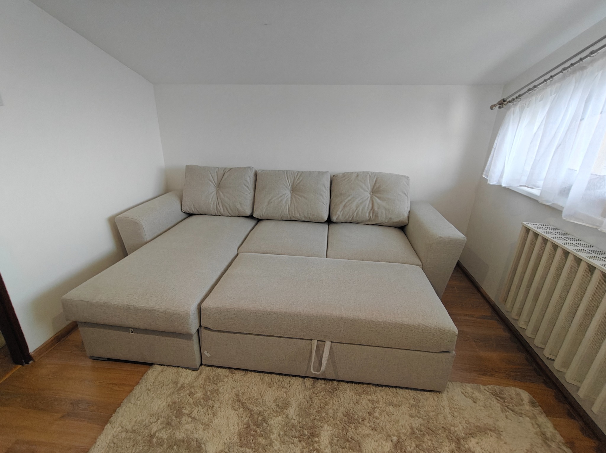 Rozłożona, beżowa sofa z poduszkami w jasnym pokoju, widok z przodu. Na podłodze dywan shaggy. Grzejnik pod oknem z białymi firankami.