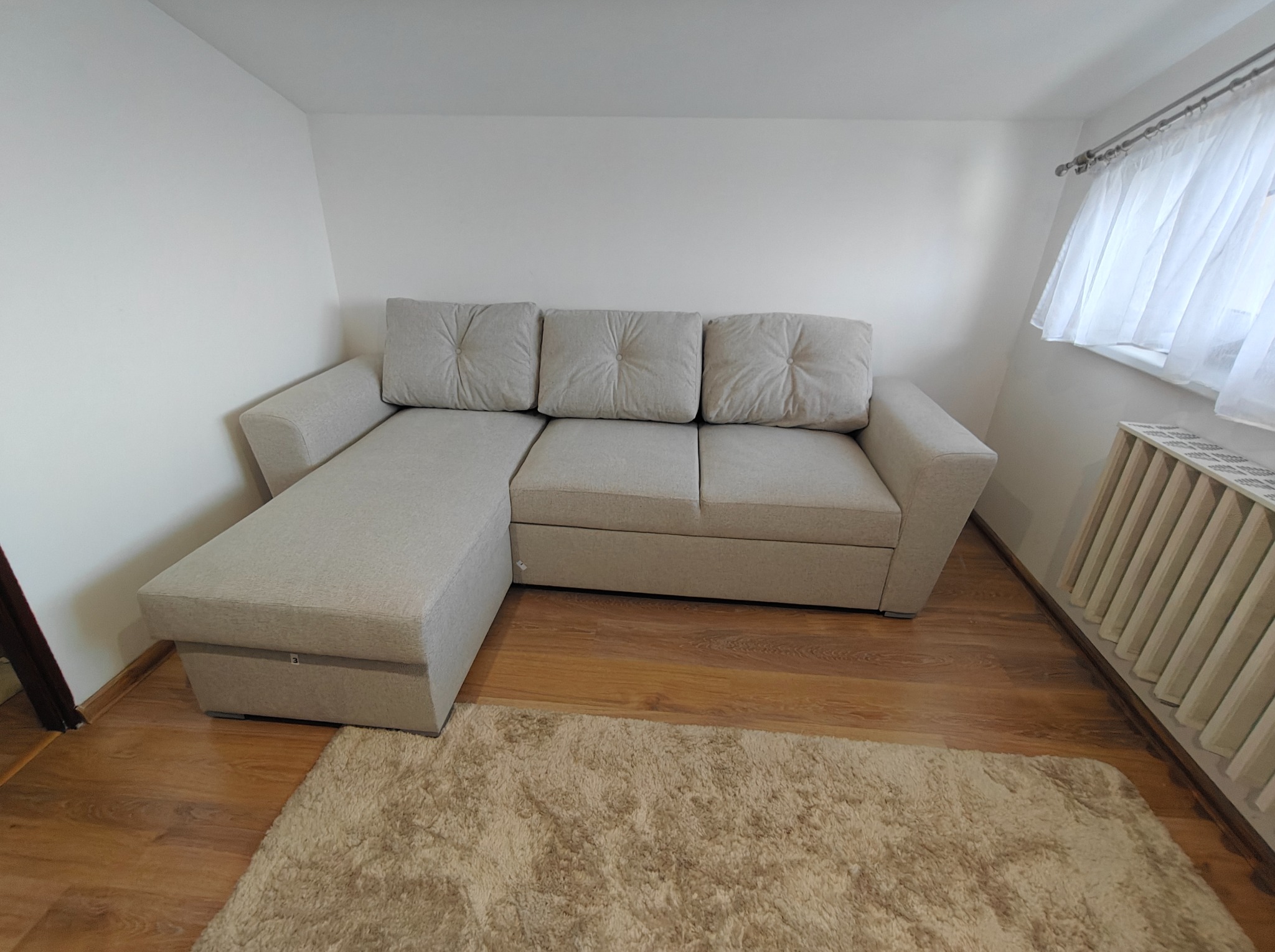 Jasny, narożny tapicerowany wypoczynek z poduszkami na drewnianej podłodze z beżowym dywanem. W tle biała ściana i grzejnik pod oknem z białymi zasłonami.