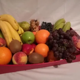 Kosz czerwony wypełniony różnorodnymi owocami: bananami, gruszkami, jabłkiem, pomarańczami, kiwi, nektarynkami, ciemnymi winogronami, borówkami i orzechami, częściowo w plastikowych pojemnikach.