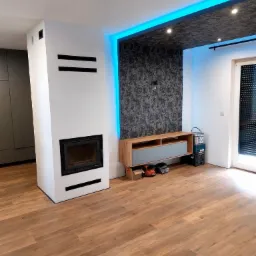 Nowoczesny salon po remoncie: drewnopodobna podłoga, biały kominek z czarnymi akcentami, ściana z efektem 3D i podświetleniem LED. Widoczne akcesoria malarskie.