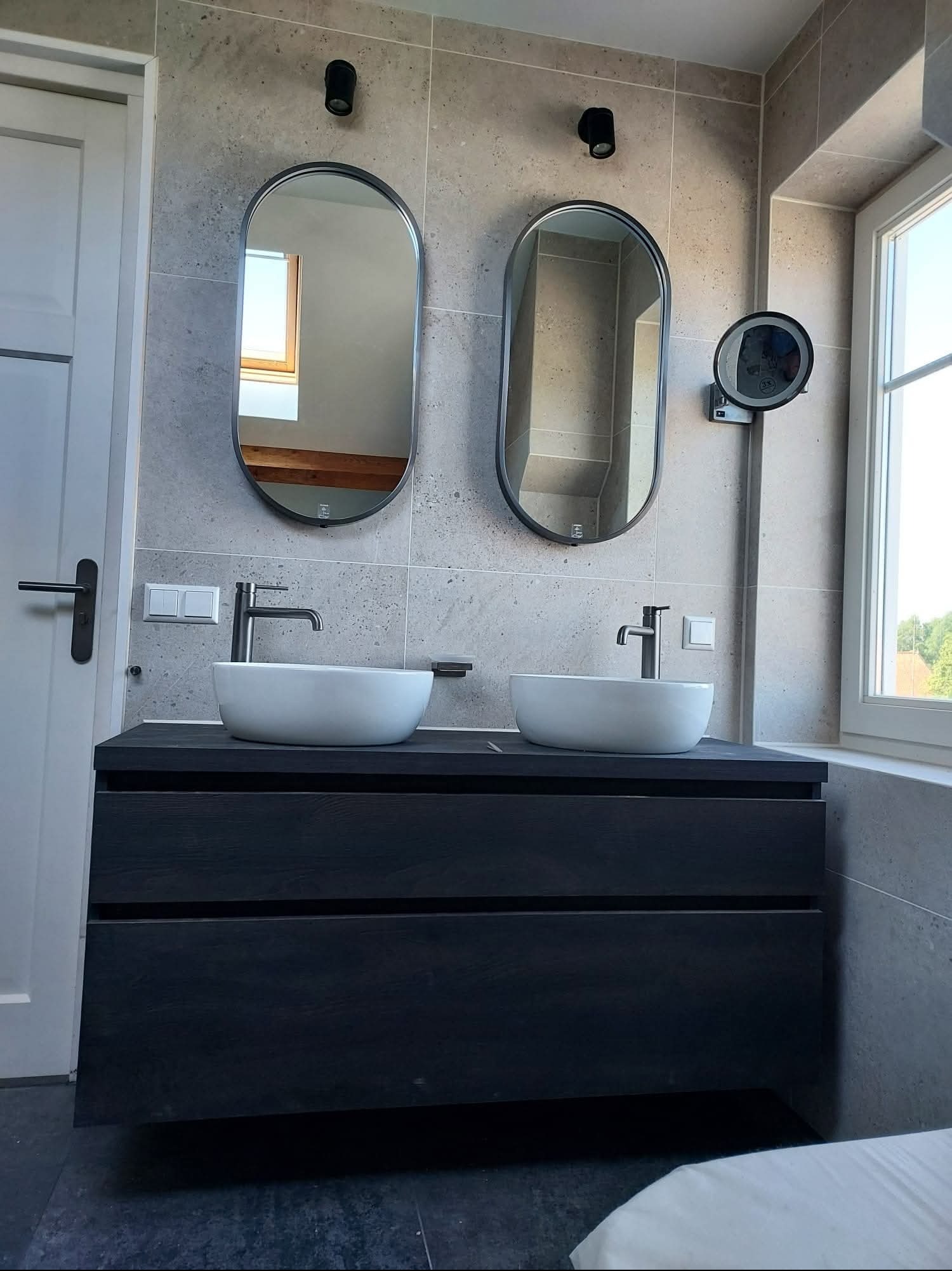 Nowoczesna łazienka z dwiema owalnymi umywalkami nablatowymi, czarną szafką pod umywalką i lustrami w czarnych ramach na tle szarych płytek.