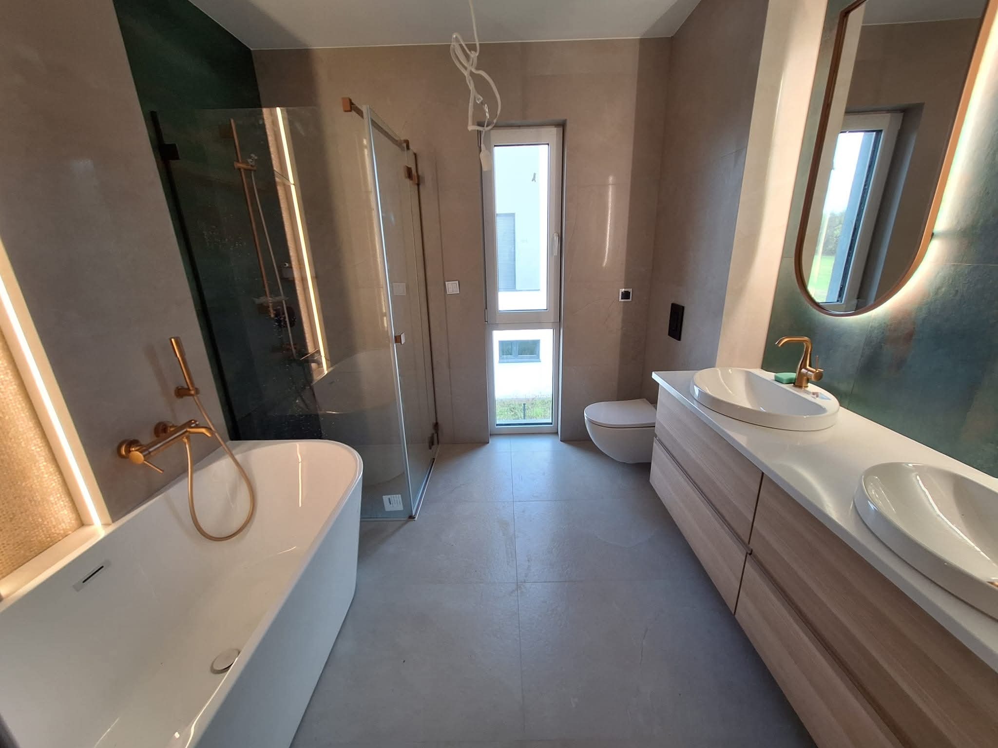 Nowoczesna łazienka z kabiną prysznicową, wanną i umywalkami nablatowymi. Wykończenie w beżach i zieleni, złota armatura, podświetlane lustro.