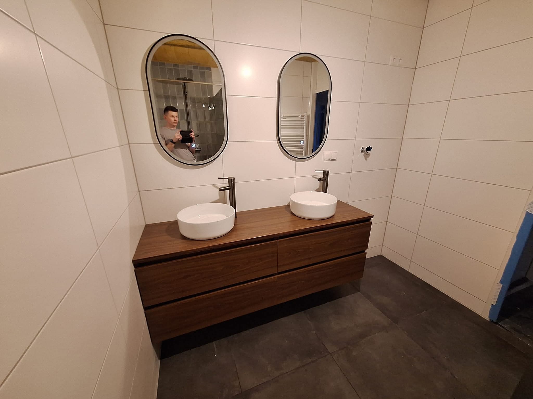 Nowoczesna łazienka z dwoma owalnymi lustrami i umywalkami nablatowymi na drewnianej szafce. W lustrze odbija się fotograf robiący zdjęcie.