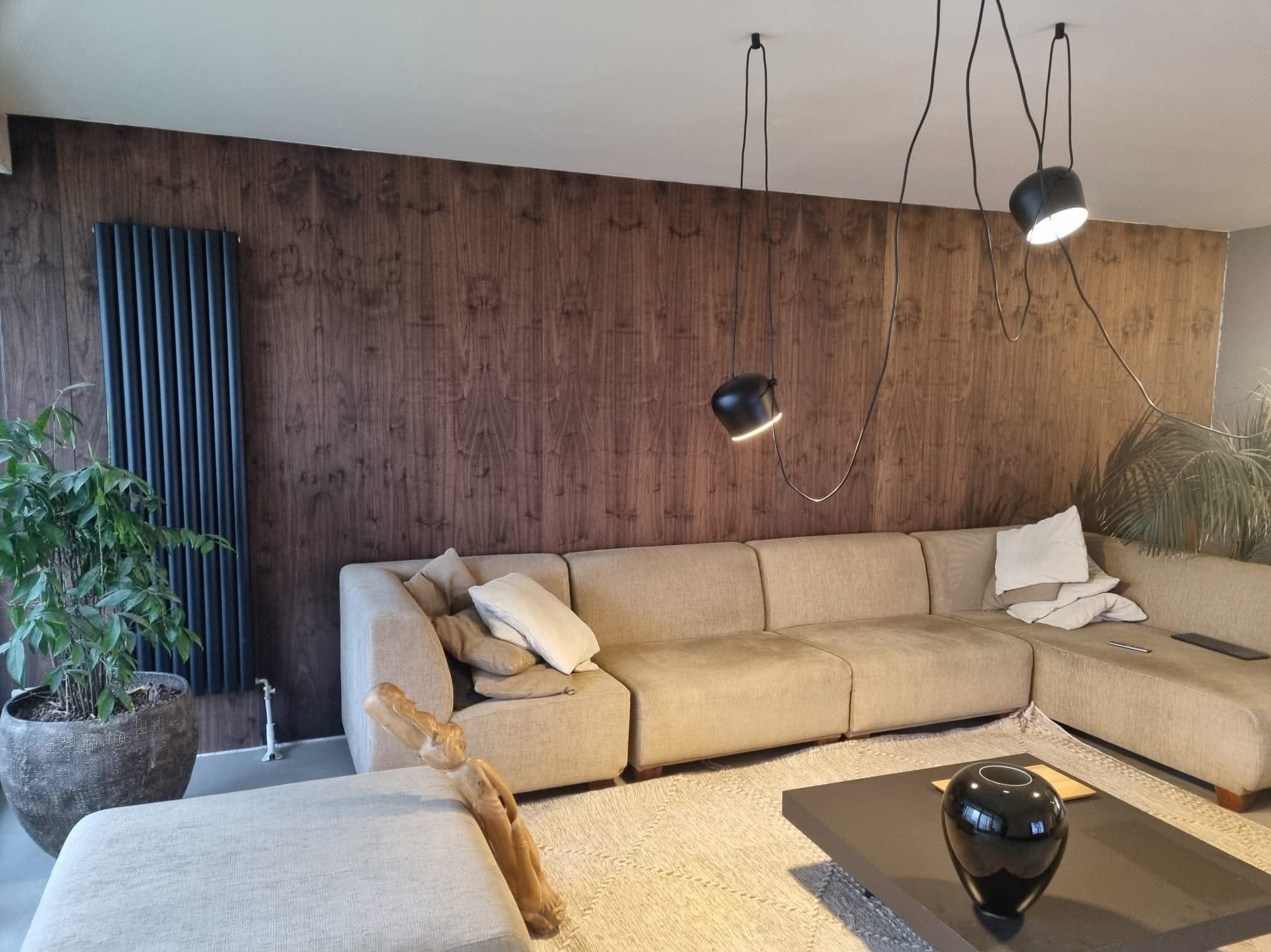 Nowoczesny salon z drewnianą ścianą, beżową sofą, czarnym grzejnikiem i designerskimi lampami. Roślina w donicy dodaje naturalnego akcentu.