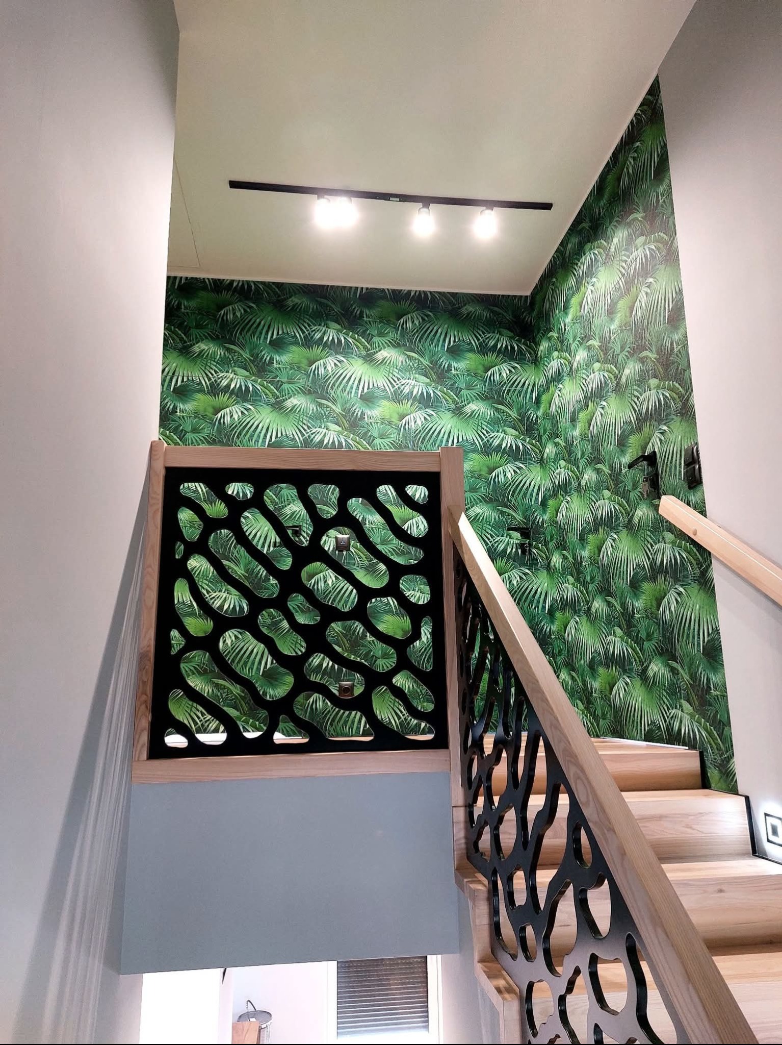 Schody z drewnianymi stopniami i czarną, ażurową balustradą. Ściana pokryta tapetą w tropikalne liście. Oświetlenie sufitowe punktowe.