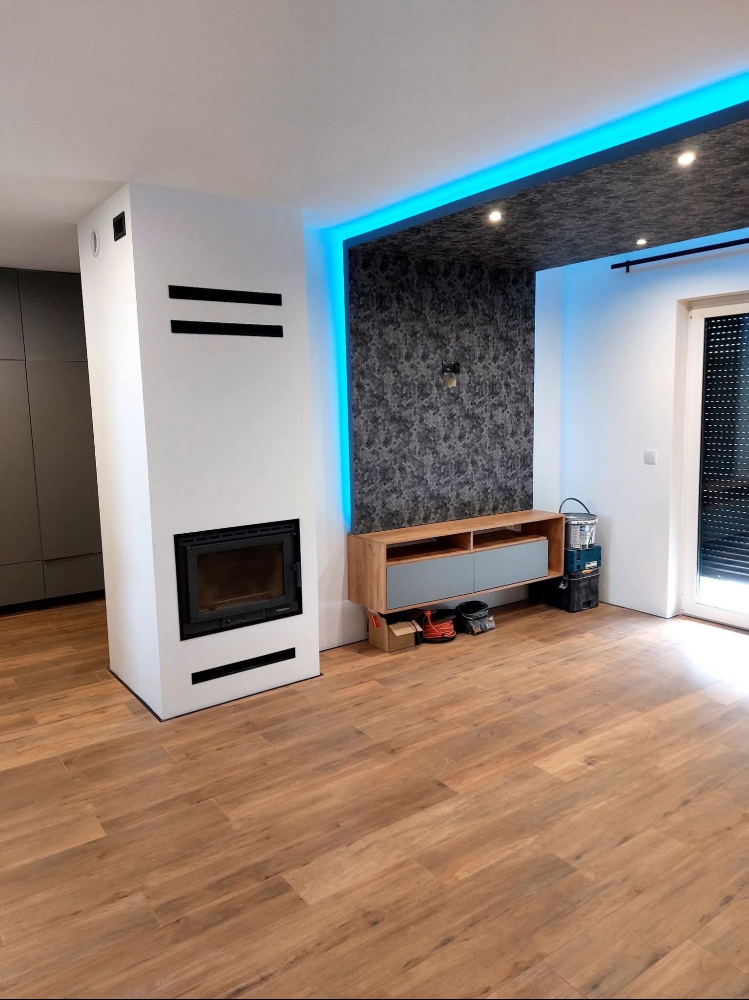 Nowoczesny salon po remoncie: drewnopodobna podłoga, biały kominek z czarnymi akcentami, ściana z efektem 3D i podświetleniem LED. Widoczne akcesoria malarskie.
