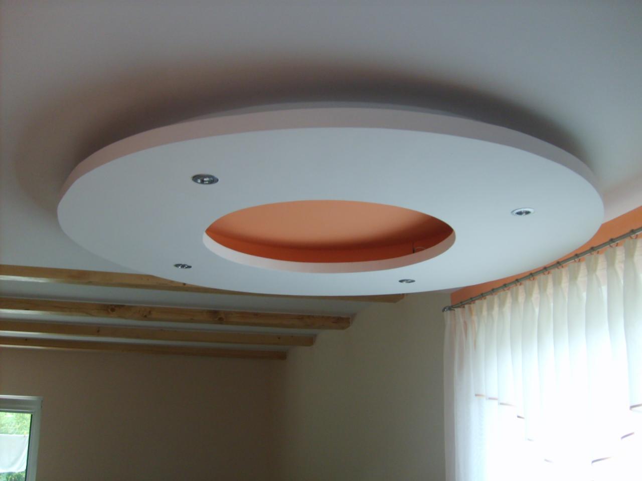 Okrągła, podwieszana konstrukcja sufitowa z oświetleniem punktowym i pomarańczowym wnętrzem, widoczna z dołu, z fragmentem drewnianej konstrukcji pod sufitem i zasłoniętym oknem w tle.