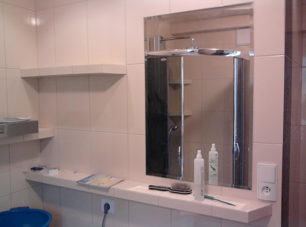 Wykończona łazienka z beżowymi płytkami, lustrem w chromowanej ramie, półkami i kabiną prysznicową widoczną w odbiciu lustra. Na blacie pod lustrem szczotka do włosów, kosmetyki i gniazdko...