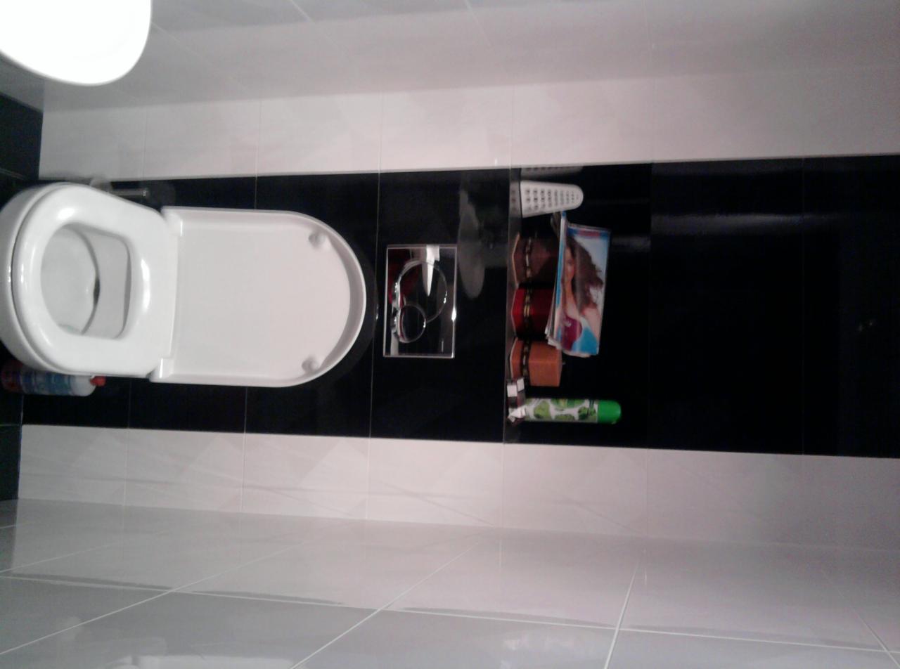 Biała toaleta z deską w pozycji otwartej na tle czarno-białych płytek, widoczny przycisk spłuczki i półka z akcesoriami łazienkowymi.