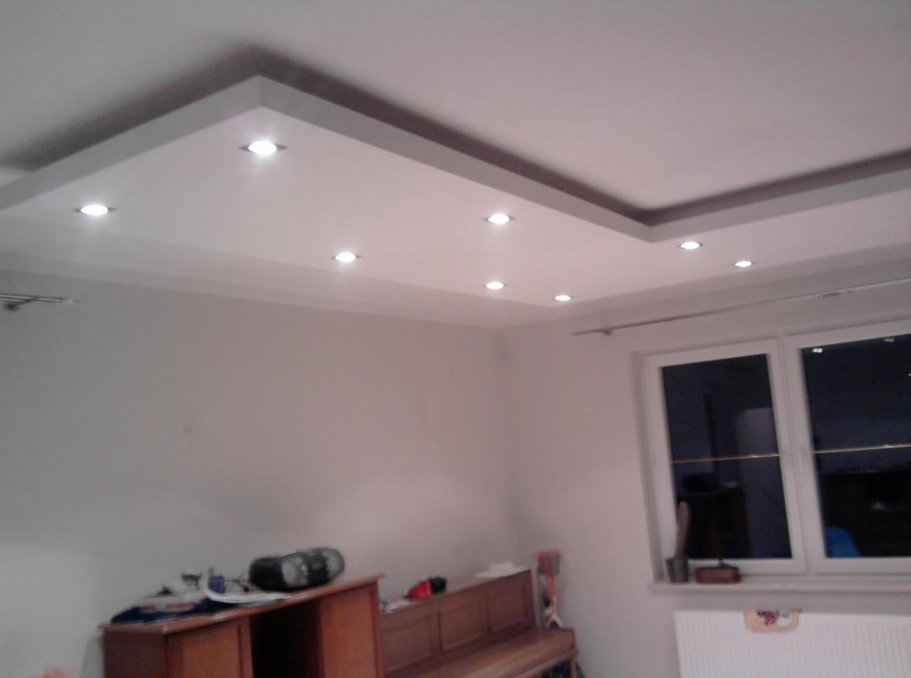 Nowoczesny sufit podwieszany z oświetleniem punktowym w kształcie prostokąta, szara listwa ozdobna, widok z dołu, fragment ściany i okna.