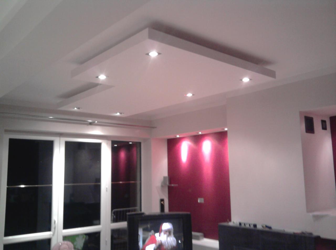 Nowoczesny sufit podwieszany z oświetleniem punktowym w salonie z oknami i ścianą akcentową w odcieniach różu, widoczne fragmenty telewizora.