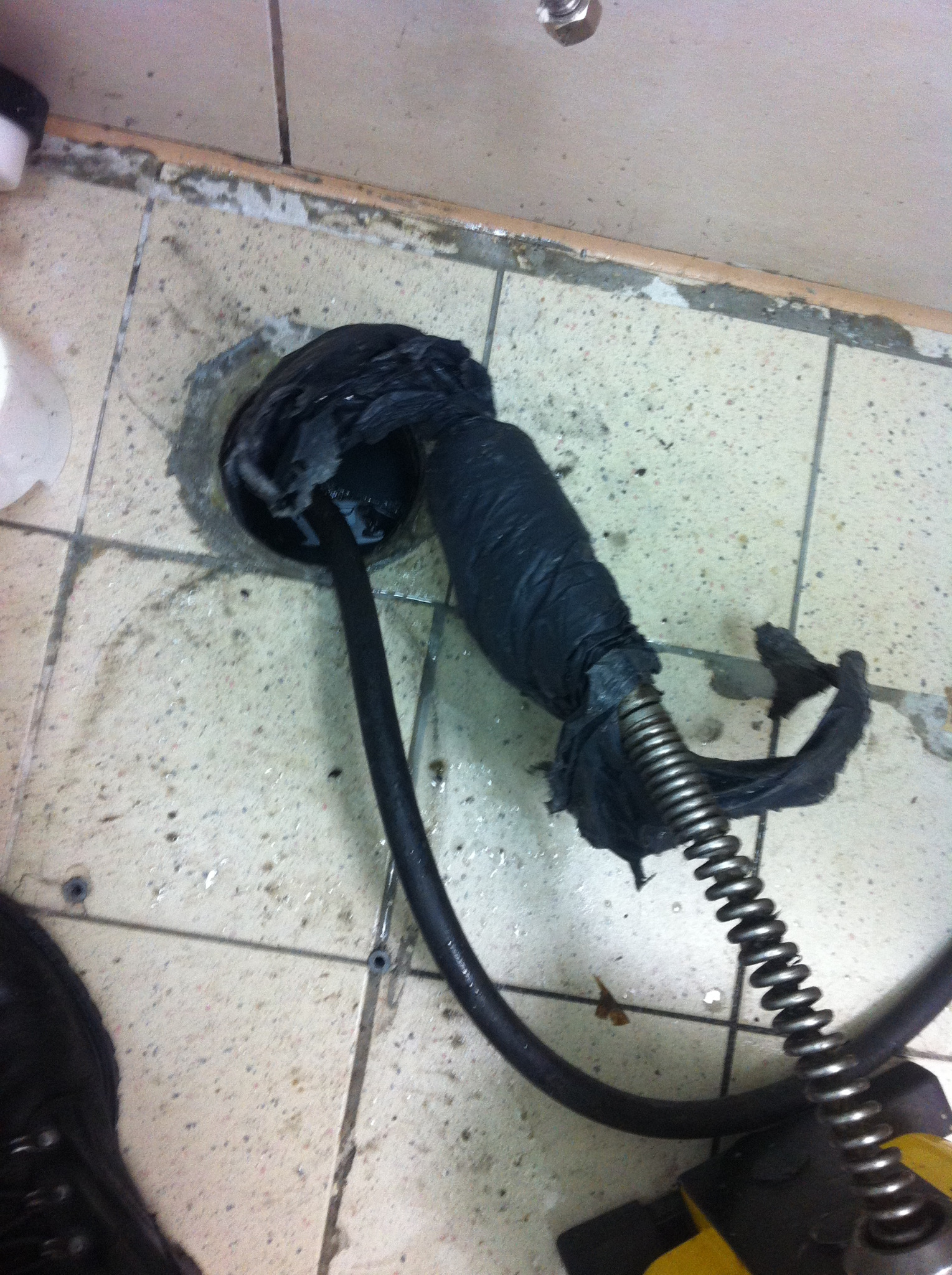 Sprężyna do udrażniania rur w otworze odpływowym na brudnej, wykafelkowanej podłodze, otoczona resztkami zatoru i wodą.