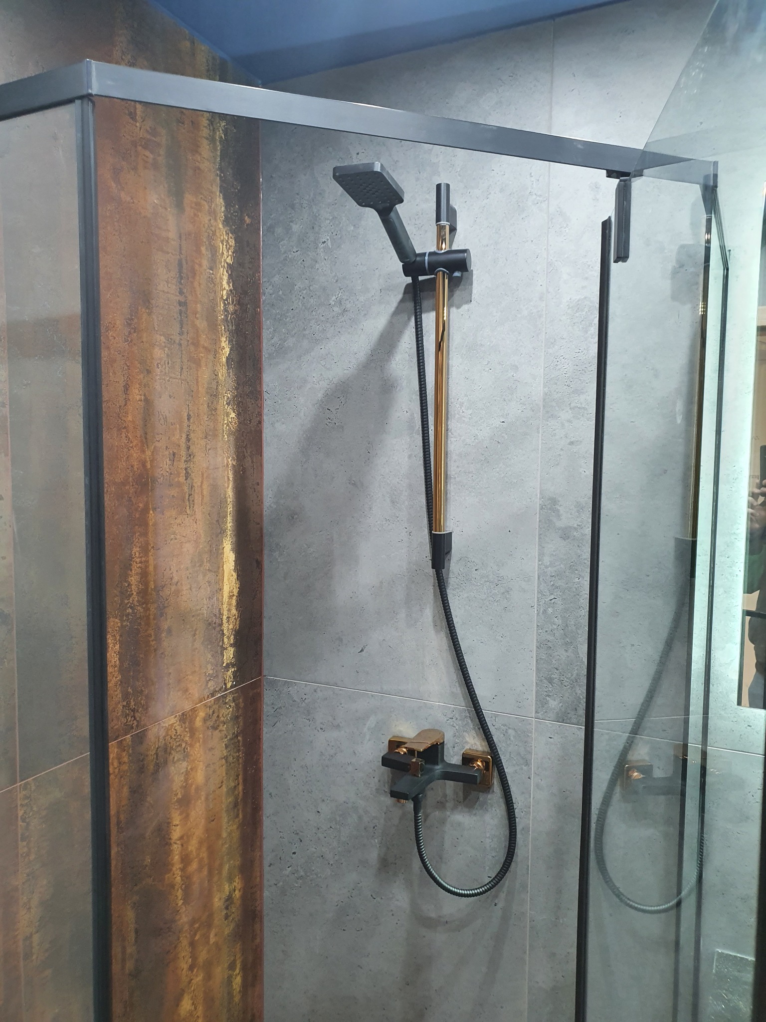Nowoczesna kabina prysznicowa z czarną armaturą z elementami w kolorze złota, szare płytki oraz rustykalne akcenty na ścianie.