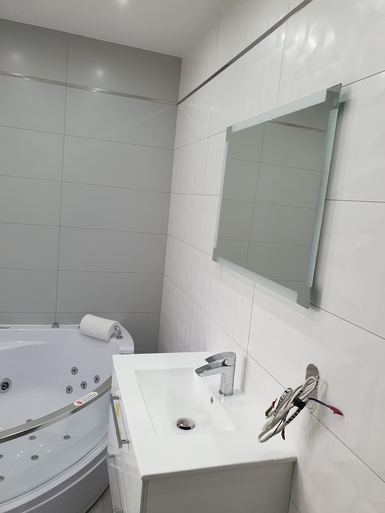 Nowoczesna łazienka z białą armaturą: narożna wanna z hydromasażem, umywalka z szafką, lustro bez ramy i wystające kable instalacji elektrycznej.