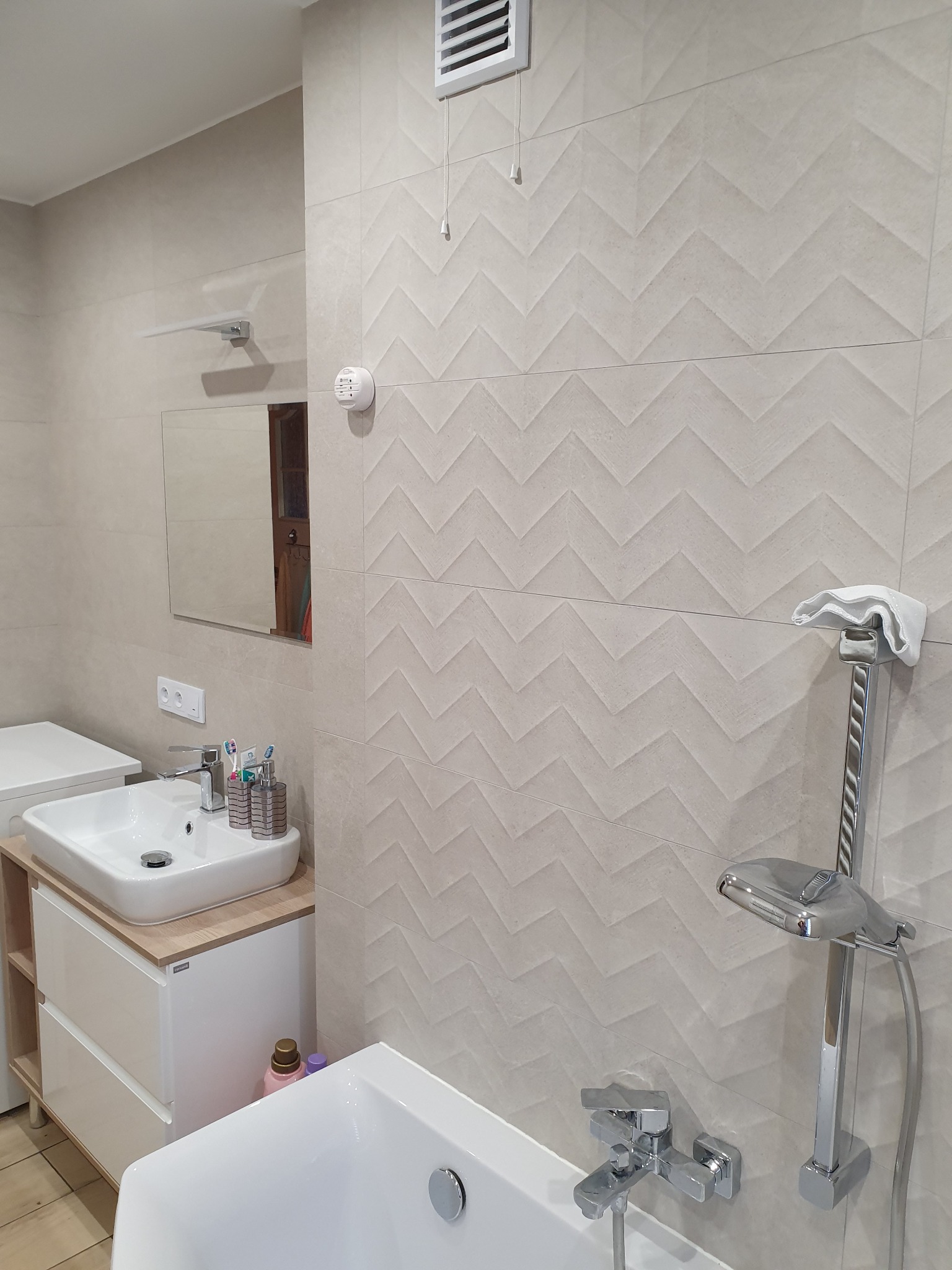 Jasna łazienka z umywalką na szafce z drewnianym blatem, białą wanną, panelami ściennymi z wzorem w jodełkę, chromowaną armaturą i akcesoriami.