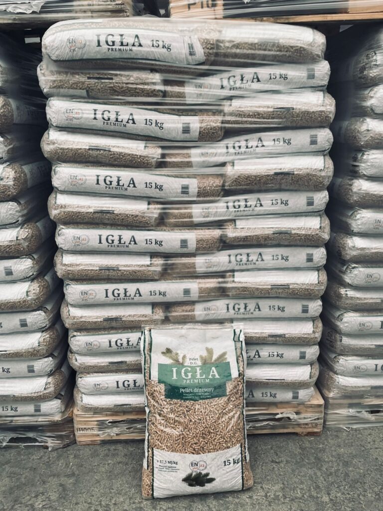 Stos worków pelletu drzewnego marki Igła Premium 15 kg, ułożonych na paletach. Na pierwszym planie pojedynczy worek pelletu. Widoczne oznaczenia certyfikacyjne.