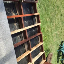 Konstrukcja drewnianego tarasu w trakcie budowy, częściowo pomalowana bejcą, widoczna puszka z bejcą i pędzlem, czarna folia pod konstrukcją, trawa w tle.