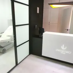 Wnętrze prywatnej kliniki z recepcją z logo 'Lewandowicz clinique' i gabinetem zabiegowym z fotelem kosmetycznym widocznym za przeszklonymi drzwiami.