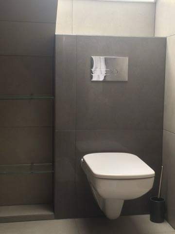 Nowoczesna toaleta podwieszana z szarymi płytkami, chromowany panel spłukujący i szklane półki w minimalistycznej łazience.