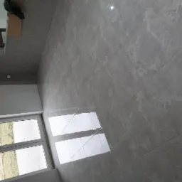 Ukończona podłoga wyłożona szarymi płytkami imitującymi marmur, widoczna z perspektywy narożnika pokoju z oknem i fragmentem innego pomieszczenia z wykładziną dywanową.