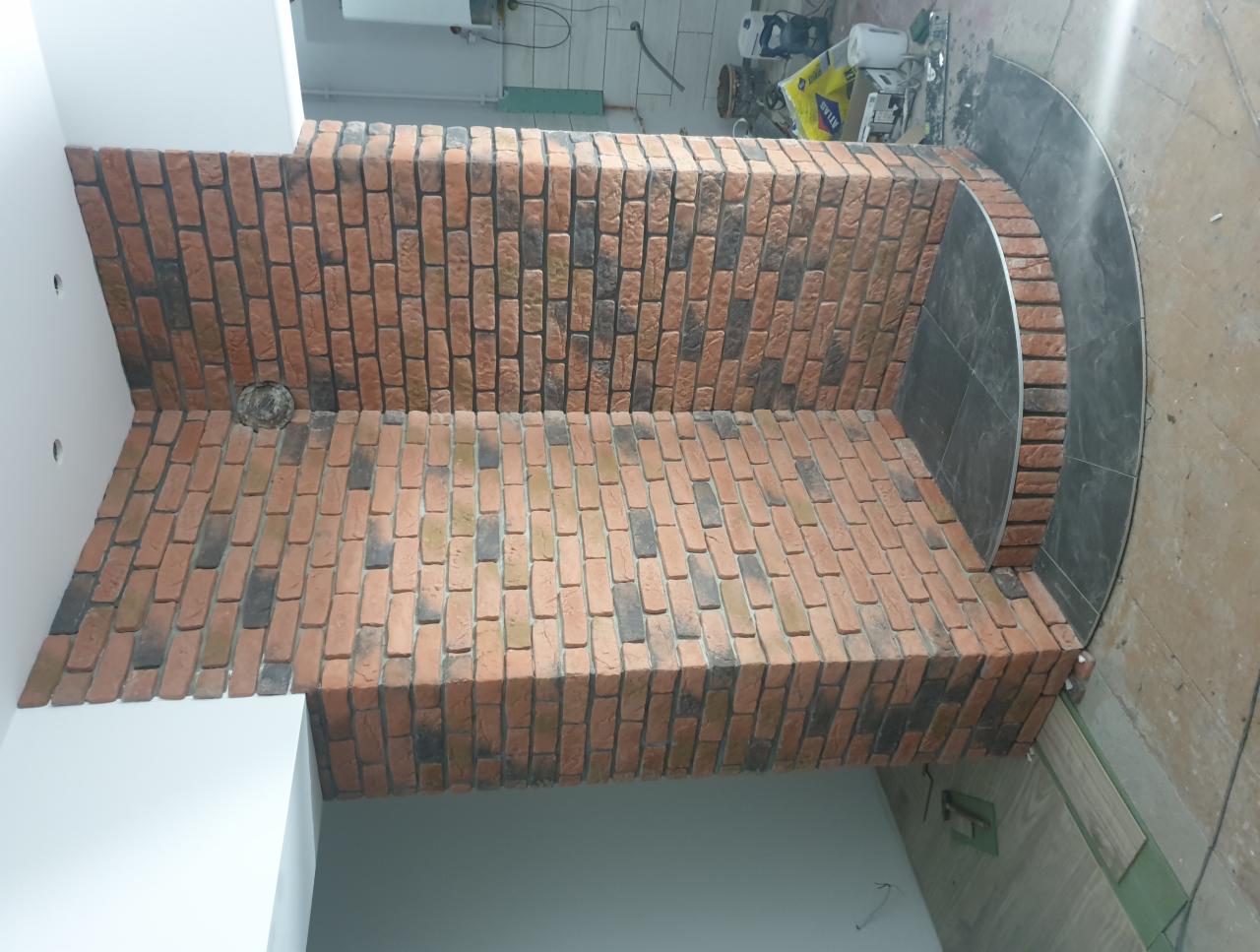 Wykończenie ściany imitacją cegły w odcieniach czerwieni i czerni, z zaokrąglonym fragmentem wyłożonym ciemnymi płytkami, widoczne narzędzia i materiały budowlane w tle.