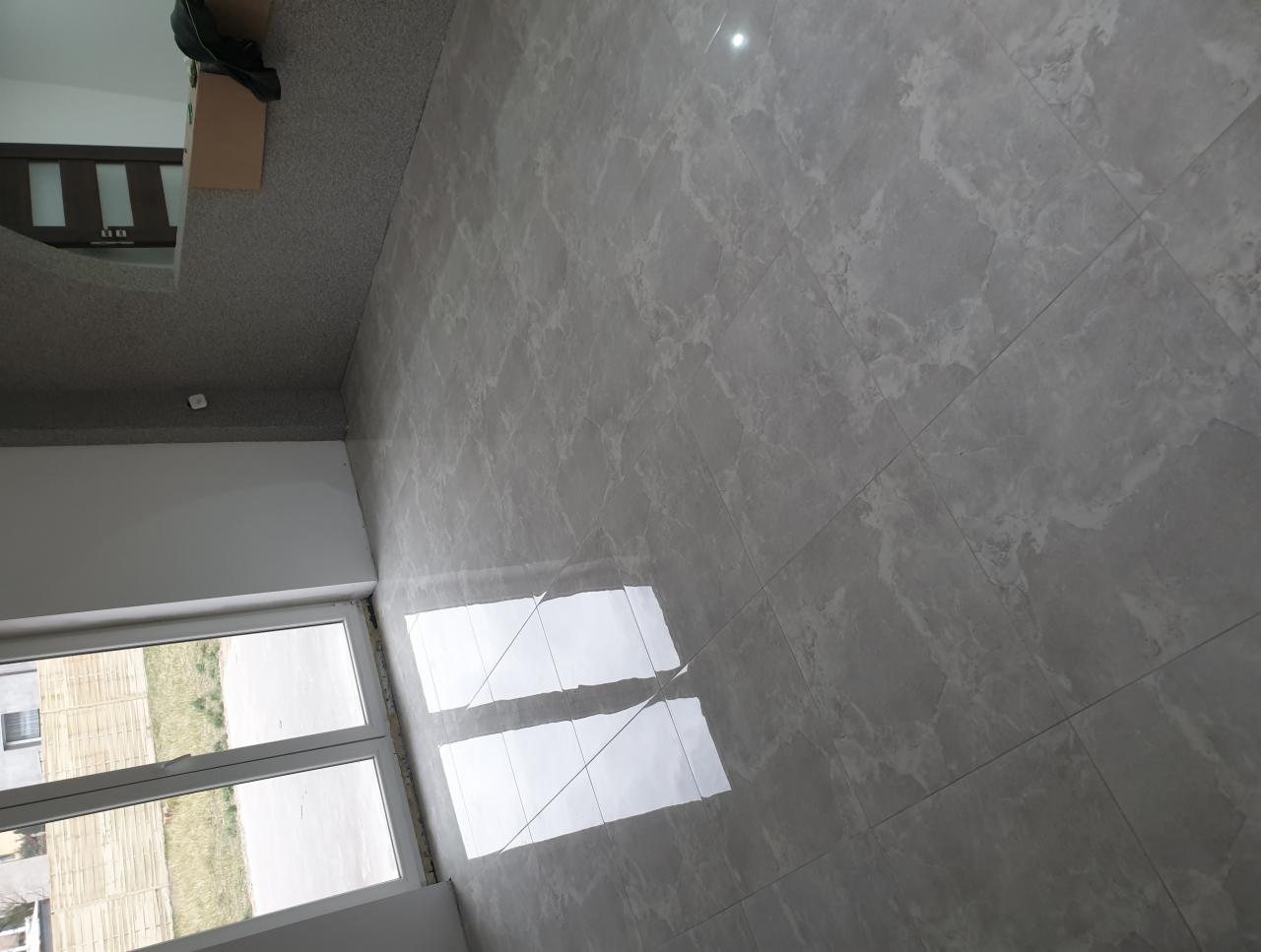 Ukończona podłoga wyłożona szarymi płytkami imitującymi marmur, widoczna z perspektywy narożnika pokoju z oknem i fragmentem innego pomieszczenia z wykładziną dywanową.