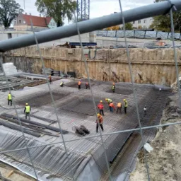 Prace zbrojarskie na placu budowy, widok przez ogrodzenie; robotnicy w kaskach i kamizelkach układają siatkę z prętów na folii, w tle wykop i żuraw budowlany.